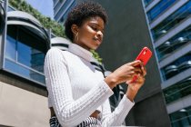 Baixo ângulo de jovem mulher negra em roupas elegantes sentado no banco e celular de navegação no dia ensolarado na rua moderna da cidade — Fotografia de Stock