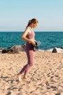 Ganzkörper-Seitenansicht einer jungen Frau in Sportbekleidung, die eine Yogamatte auf Sand legt, während sie sich auf das Training am Strand in der Nähe des Ozeans vorbereitet — Stockfoto