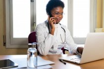 Зручний молодий афроамериканець - медик відповідає на телефонний дзвінок і користується ноутбуком, а пацієнти дистанційно консультуються з офісом. — стокове фото