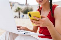 Обрізана невпізнавана фрілансерка, яка сидить на березі моря і друкує на ноутбуці і переглядає на смартфоні, працюючи віддалено на проекті влітку — стокове фото