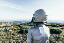 Back view male astronaut in spacesuit and helmet standing on grass and stones in highlands — Stock Photo