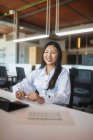 Позитивная азиатка-работница, сидящая за столом на рабочем месте и смотрящая в камеру — стоковое фото