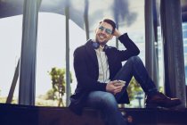 Content male in stylish apparel with headphones and cellphone sitting in city on sunny day — Stock Photo