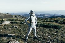 Astronauta masculino de cuerpo completo con traje espacial y casco cogido de la mano en la cintura mientras está de pie sobre hierba y piedras en las tierras altas - foto de stock