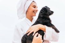 Ottimista giovane donna in accappatoio e asciugamano sorridente e abbracciare cane nero durante la sessione spa a casa — Foto stock