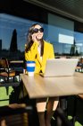 Усміхнена азіатська бізнес-леді з жовтим пальто, сидячи за столом, має каву зі своїм смартфоном і ноутбуком — стокове фото