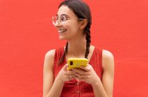 Fröhliche junge Frau mit Zöpfchenfrisur surft auf dem Smartphone und schaut auf rotem Hintergrund auf der Straße weg — Stockfoto