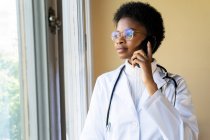 Junge afroamerikanische Ärztin in weißem Mantel mit Stethoskop spricht Handy, während sie in der Klinik am Fenster steht — Stockfoto