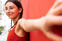 Seitenansicht einer zufriedenen Frau mit trendiger Brille, die sich an eine rote Hauswand in der Straße lehnt und mit ausgestrecktem Arm in die Kamera blickt — Stockfoto