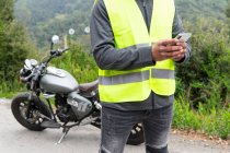 Обрезание мужской байкер в жилете сообщения на мобильный телефон, стоя рядом со сломанным мотоциклом возле пышных зеленых лесов — стоковое фото