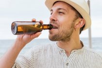 Щасливий молодий бородатий хіпстер чоловік мандрівник у стильній літній сорочці та капелюсі насолоджується холодним пивом з пляшки під час охолодження на узбережжі під час літніх канікул — стокове фото