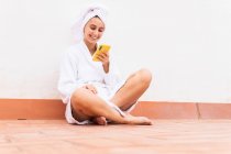 Mujer joven en albornoz y toalla sonriendo y navegando por el teléfono móvil mientras descansa en el balcón después de la ducha - foto de stock