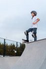 Teenage skater in protective gear riding skateboard during weekend in skate park and looking away — Stock Photo