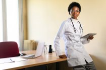 Позитивний молода чорна жінка-лікар в медичному пальто і окулярах зі стетоскопом дивиться на камеру під час роботи з планшетом в сучасному клінічному офісі — стокове фото