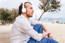 Vista lateral de un joven barbudo despreocupado con una elegante camisa casual escuchando música a través de auriculares inalámbricos y disfrutando de la brisa fresca mientras pasa el día de verano en la playa de arena cerca del mar - foto de stock