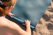 Зверху обрізана невизначена молода жінка в спортивному одязі розміщує йога килимок на піску, готуючись до практики на пляжі поблизу океану — стокове фото