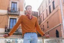 Снизу молодой кудрявый парень в стильной разноцветной полосатой рубашке стоит на улице и задумчиво смотрит в камеру. — стоковое фото