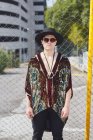 Уверенный мужчина в модных солнцезащитных очках и шляпе стоит рядом с сеткой забора на улице и смотрит в камеру — стоковое фото