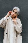 Serious transgender female in trendy coat leaning on gray wall while standing in street looking at camera — Stock Photo