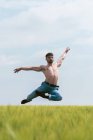 Мужчина без рубашки в джинсе совершает чувственный балетный прыжок с руками, раскинутыми над высокой травой на мрачном поле — стоковое фото