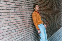 Jovem elegante pensativo étnico encaracolado cara na roupa da moda inclinando-se contra a parede de tijolo na rua urbana olhando para a câmera — Fotografia de Stock