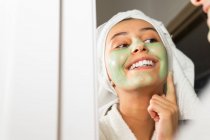 Счастливая женщина с игрушкой на голове улыбается и наносит зеленую маску на лицо, глядя в зеркало в ванной комнате дома — стоковое фото