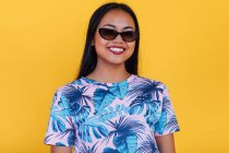 Mulher asiática feliz em óculos de sol elegantes e t-shirt com impressão de folha tropical olhando para a câmera no fundo amarelo no estúdio — Fotografia de Stock