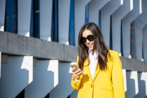 Femme d'affaires asiatique avec manteau jaune et téléphone intelligent marchant dans la rue avec bâtiment en arrière-plan — Photo de stock