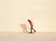 Vue latérale du skateboarder masculin avec les mains dans les poches regardant loin sur la chaussée à la lumière du soleil sur fond beige — Photo de stock