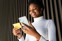 De baixo positivo jovem afro-americano feminino em roupas da moda sorrindo e usando cartão de crédito e smartphone para pagar a ordem on-line contra a parede listrada na rua — Fotografia de Stock