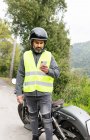 Задумчивый взрослый латиноамериканец-байкер в защитном шлеме и жилете, отправляющий сообщения по мобильному телефону, стоя рядом со сломанным мотоциклом возле пышных зеленых лесов — стоковое фото