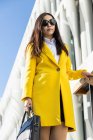Asian business woman with yellow coat and smart phone walking on the street with building in the background — Stock Photo