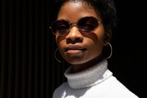 Mujer afroamericana joven en suéter elegante y gafas de sol mirando a la cámara mientras está de pie en la luz del sol brillante contra el fondo negro - foto de stock