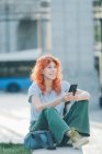 Вид сбоку веселый рыжая женщина сидит на улице и сообщения в социальных сетях на мобильном телефоне, слушая музыку — стоковое фото