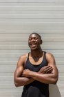 Apto afro-americano masculino de pé com braços cruzados perto de parede de metal na rua e rindo com os olhos fechados no dia ensolarado — Fotografia de Stock