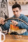 Serious male mechanic fixing wheel spoke while sitting at workbench in bike service — Stock Photo