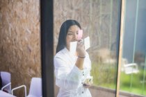 Sonriente empleada asiática escribiendo en una nota adhesiva en una pared de vidrio mientras trabaja en una oficina moderna - foto de stock
