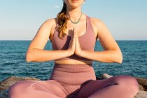 Hembra serena irreconocible recortada con los ojos cerrados haciendo yoga en pose de loto durante la meditación sobre roca en la orilla del mar - foto de stock