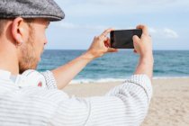 Боковой вид молодого стильного бородатого мужчины, который фотографирует мыс на мобильный телефон, проводя летний день на песчаном пляже — стоковое фото