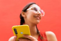 De baixo alegre jovem fêmea em pigtails penteado navegando no smartphone de pé olhando para longe no fundo vermelho na rua — Fotografia de Stock