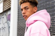Seitenansicht eines selbstbewussten jungen lockigen Hipsters im rosafarbenen Kapuzenpulli, der in die Kamera gegen eine Ziegelwand blickt — Stockfoto