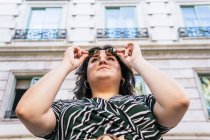 Low angle of confident young curvy female in stylish dress with geometric print and trendy sunglasses looking away while standing near stone urban building in summer day — Stock Photo