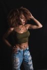 Capelli ricci donna afroamericana in crop top alla moda e jeans in piedi con mano in vita su sfondo nero in studio — Foto stock