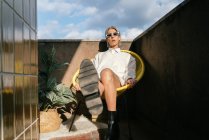 Selbstbewusste Transgender-Frau im trendigen Outfit streckt ihr Bein in die Kamera, während sie im Sessel auf der Terrasse sitzt und in die Kamera blickt — Stockfoto