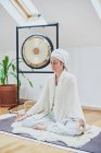 Зрелая женщина с закрытыми глазами сидит со скрещенными ногами на пушистом ковре, практикуя йогу дома — стоковое фото