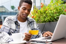 Focused African American male paying for order with plastic card while using laptop during online shopping in street cafe — Stock Photo