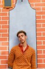 Young stylish thoughtful ethnic curly haired guy in trendy outfit leaning against brick wall on urban street looking at camera — Stock Photo