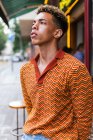 Молодий етнічний кучерявий хлопець у стильній барвистій смугастій сорочці, що стоїть на вулиці, дивлячись у відчайдушно — стокове фото