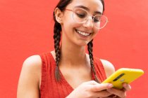 Fröhliche junge Frau mit Zöpfchenfrisur surft auf Smartphone auf rotem Hintergrund auf der Straße — Stockfoto