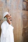 Cheerful Muslim male in traditional clothes smiling and browsing cellphone while standing near shabby wall on street — Stock Photo